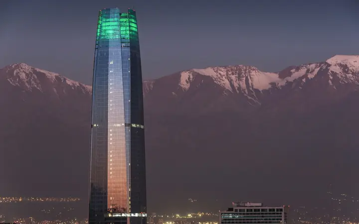 Torre de vidrio iluminada al anochecer frente a la cordillera